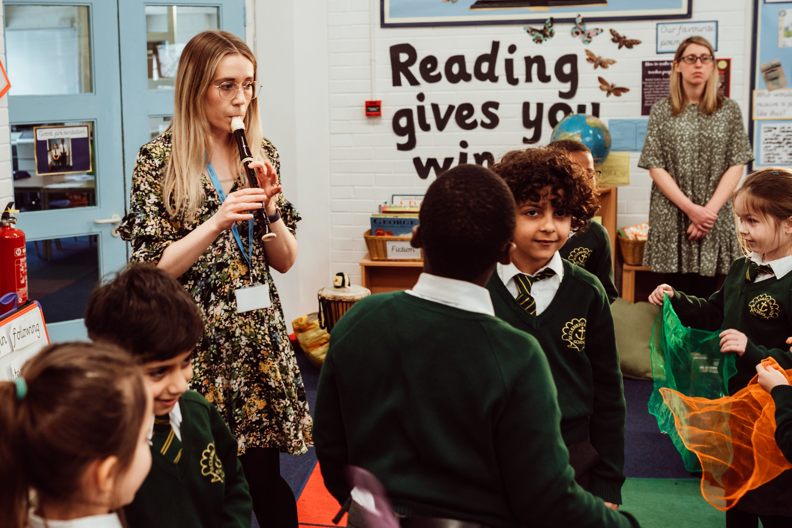 A music lesson is in full swing with a group of engaged primary school children wearing green uniforms, watching as their teacher plays the recorder. The classroom environment is vibrant and educational, with a "Reading gives you wings" display in the background, adding to the atmosphere of learning and creativity. Another teacher observes quietly, ensuring a focused and supportive learning space.