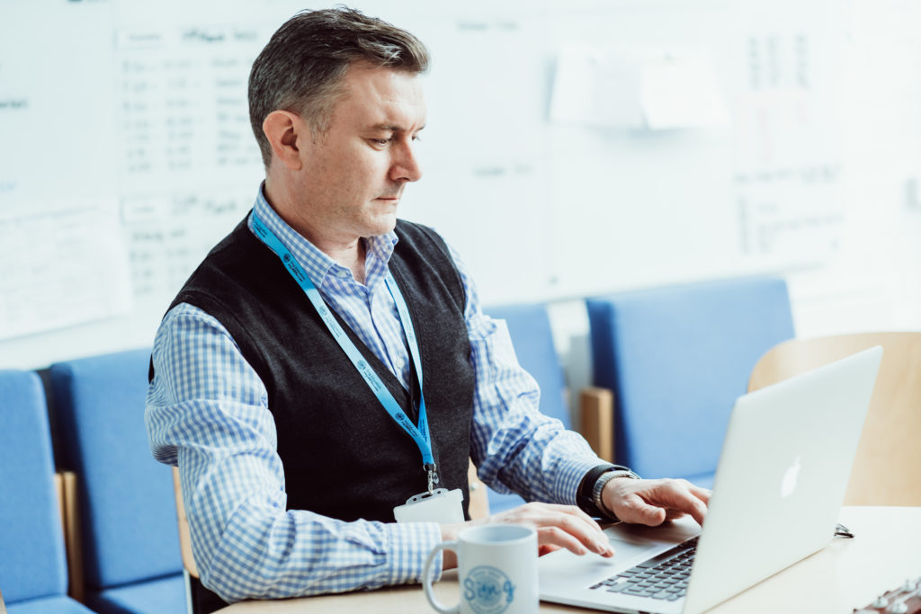A focused individual wearing a lanyard and a vest over a checkered shirt is working intently on a laptop, with a Sing Education mug nearby, suggesting a setting of professional educational planning or administration. A whiteboard with a schedule in the background indicates an organized work environment.