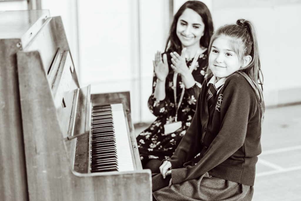 A young student sits at a piano, glancing back with a smile, while a teacher stands behind her, clapping in encouragement. The sepia tones of the photograph highlight the warmth of the moment, capturing a sense of achievement and the supportive atmosphere of the music lesson.