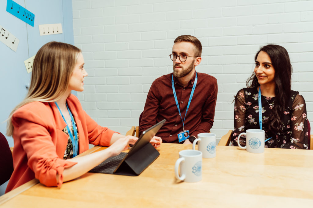 Three professionals are engaged in a collaborative meeting around a wooden table, with one person presenting on a tablet. They are in a bright room with a white brick wall adorned with musical notes, and each has a mug, suggesting a relaxed, yet focused, work environment. The lanyards they wear indicate they are part of the Sing Education team, reflecting a sense of community and shared purpose.