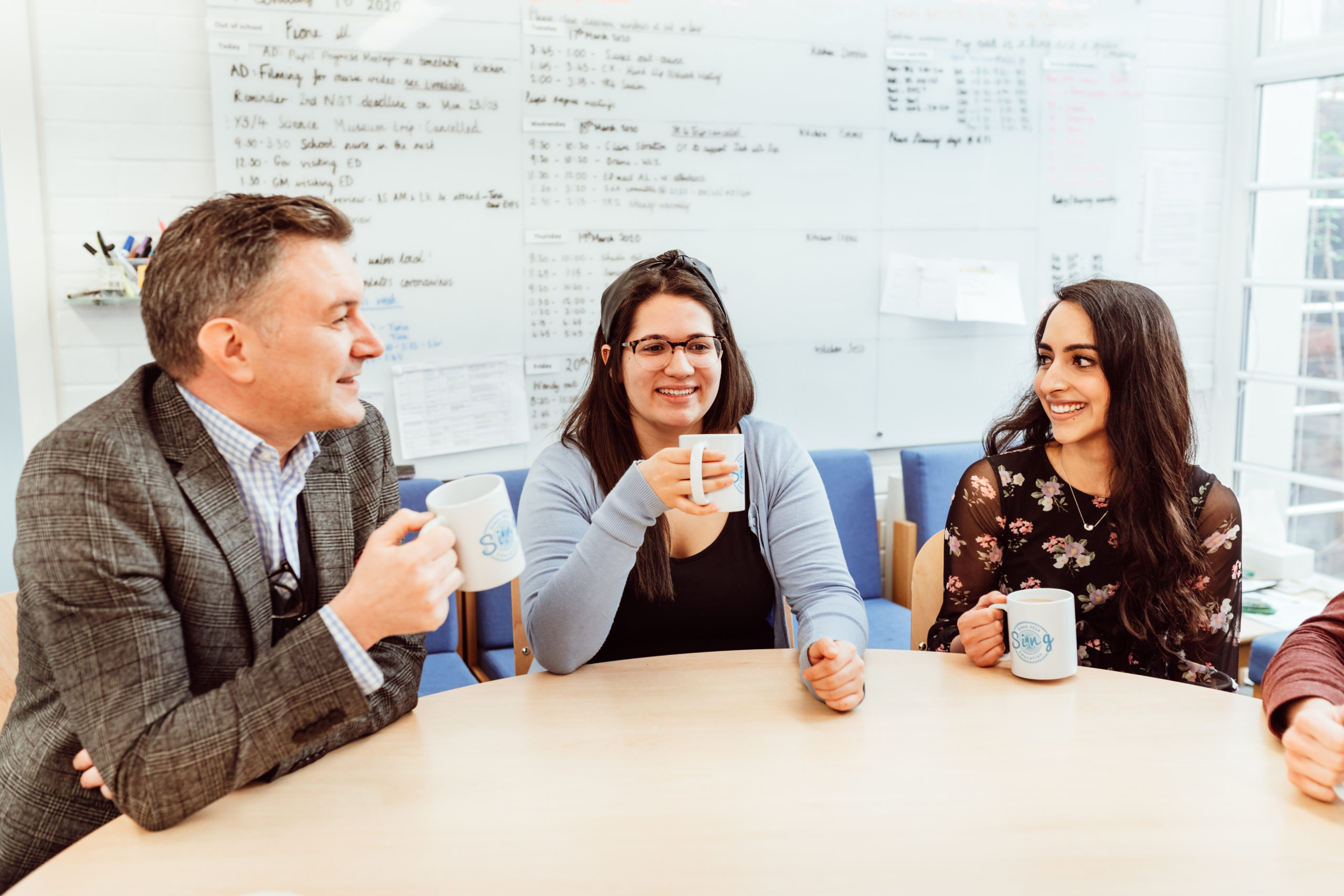 Three colleagues share a light-hearted moment around a table, each holding a mug with the Sing Education logo, suggesting a casual meeting or break. They are engaged in conversation, with expressions of enjoyment and ease, in a room with a whiteboard filled with notes and schedules, indicating a collaborative work environment.