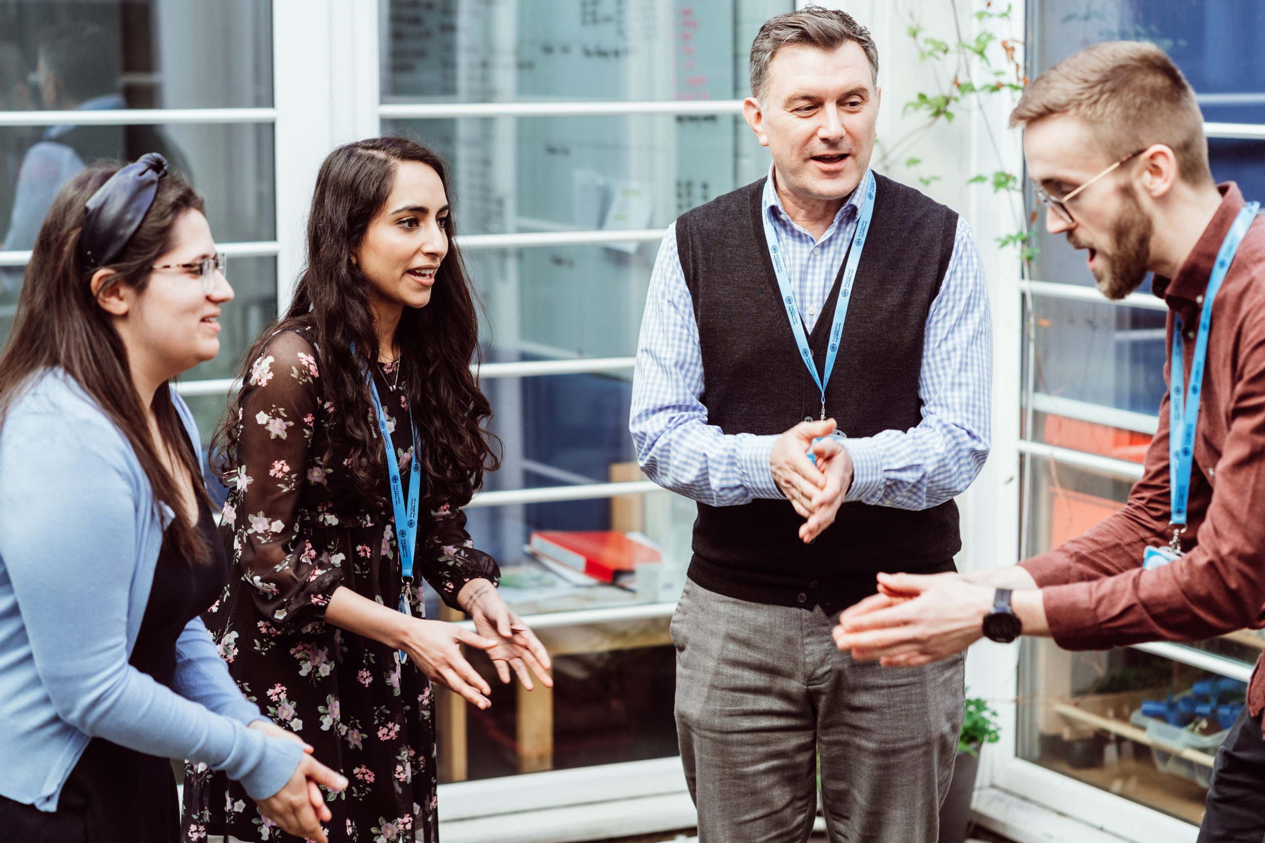 Four professionals are engaged in a lively conversation, with expressions of interest and enthusiasm. They are wearing lanyards, suggesting a work or conference setting. The atmosphere is informal and collaborative, with a backdrop that includes a window and some greenery, contributing to a relaxed yet focused exchange.