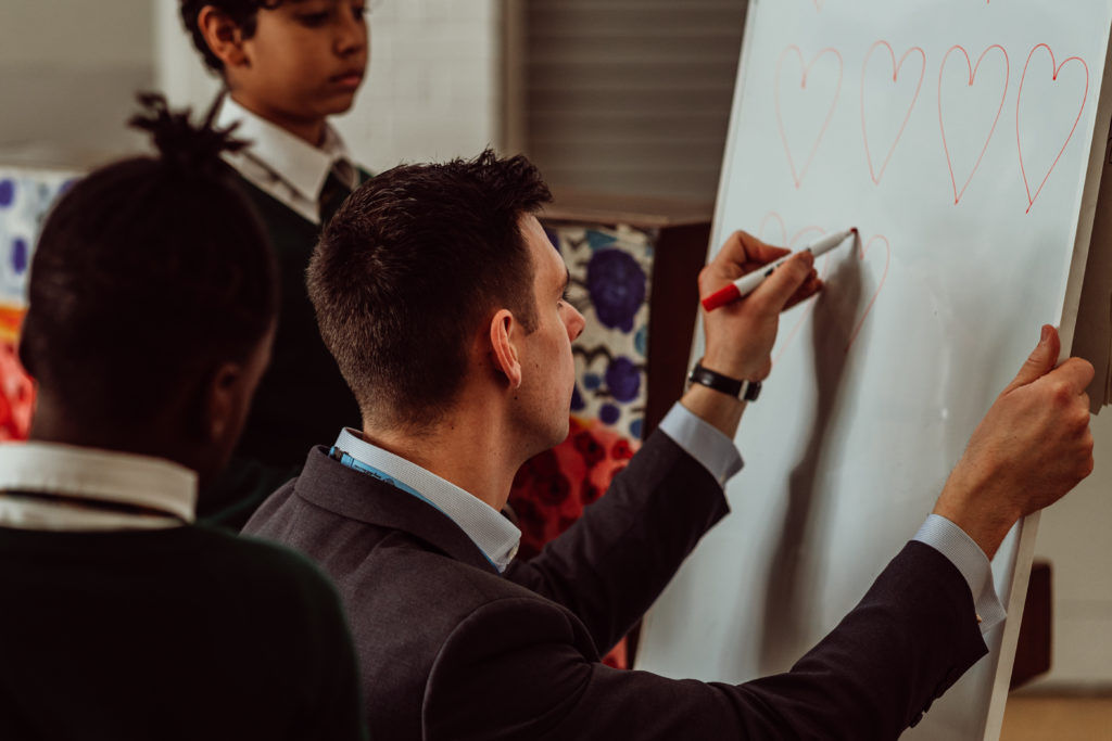 A focused teacher, with a marker in hand, draws a series of red hearts on a whiteboard, while a student attentively observes the process, suggesting a creative or illustrative lesson in a classroom setting. The atmosphere is one of concentration and learning, with the student closely following the teacher's demonstration.