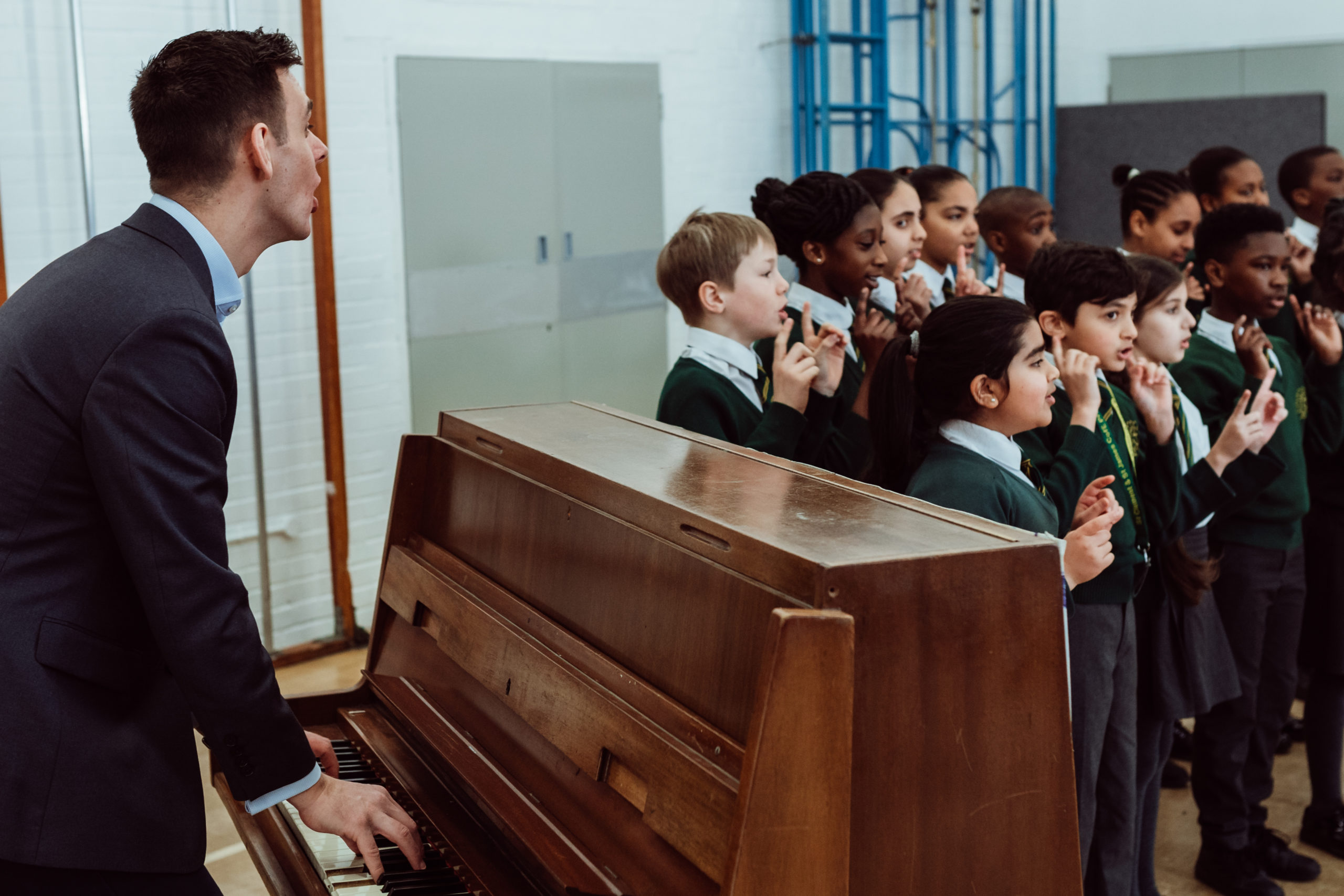 A music teacher in a suit plays a piano, facing a group of enthusiastic schoolchildren who are singing and performing hand gestures. The children, wearing green school uniforms, appear engaged and joyful as they participate in the musical activity in a spacious room with a reflective floor and gym equipment in the background.