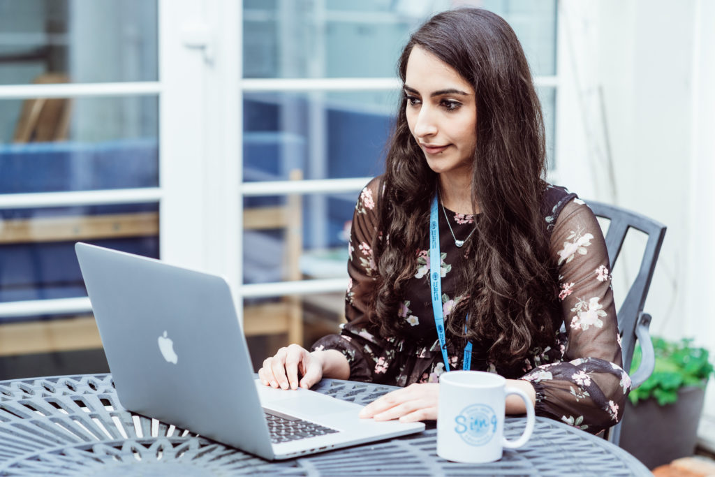 A focused woman sits at an outdoor table working on a laptop, with a Sing Education mug beside her, embodying a blend of professionalism and comfort in an informal setting.