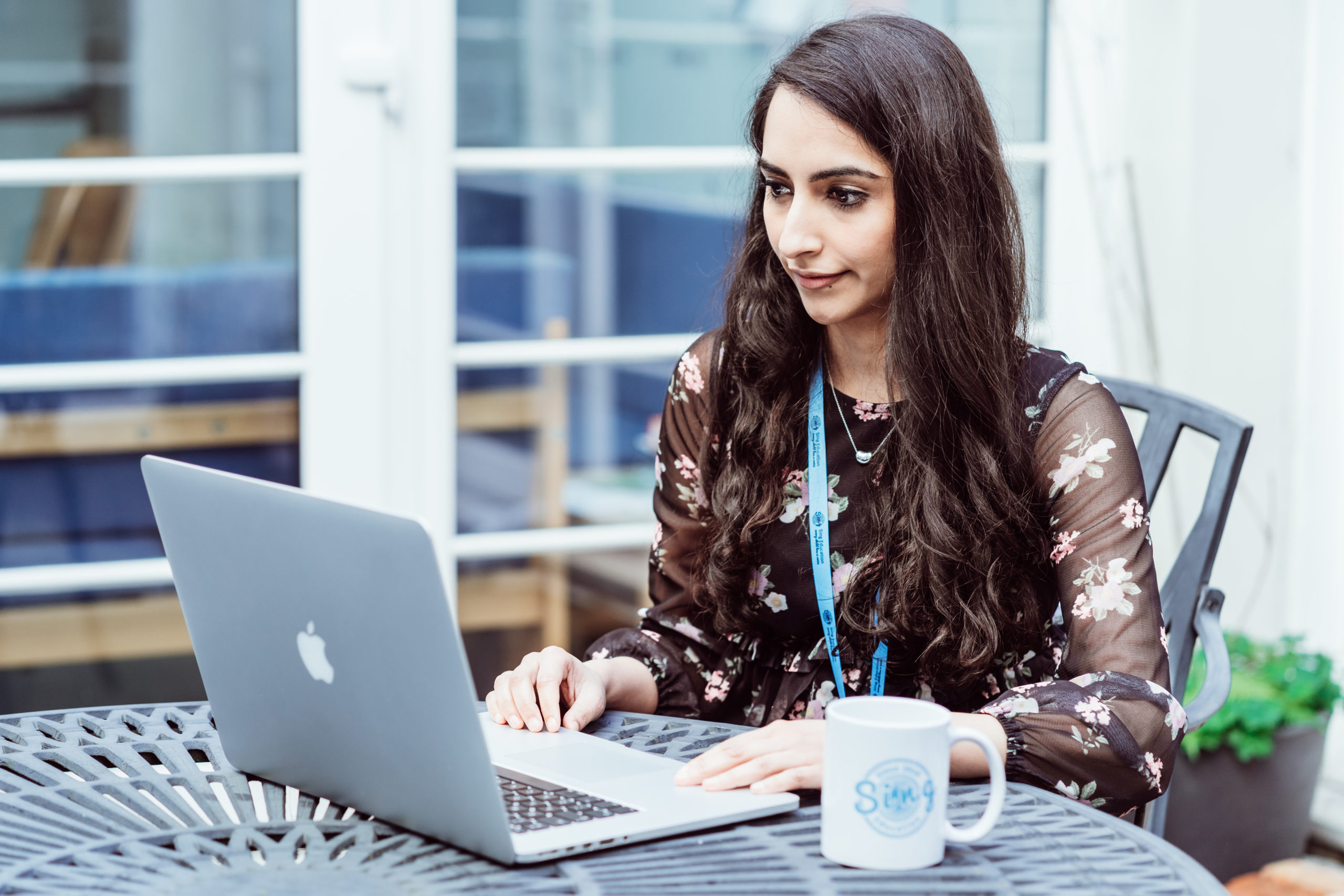 A focused woman sits at an outdoor table working on a laptop, with a Sing Education mug beside her, embodying a blend of professionalism and comfort in an informal setting.