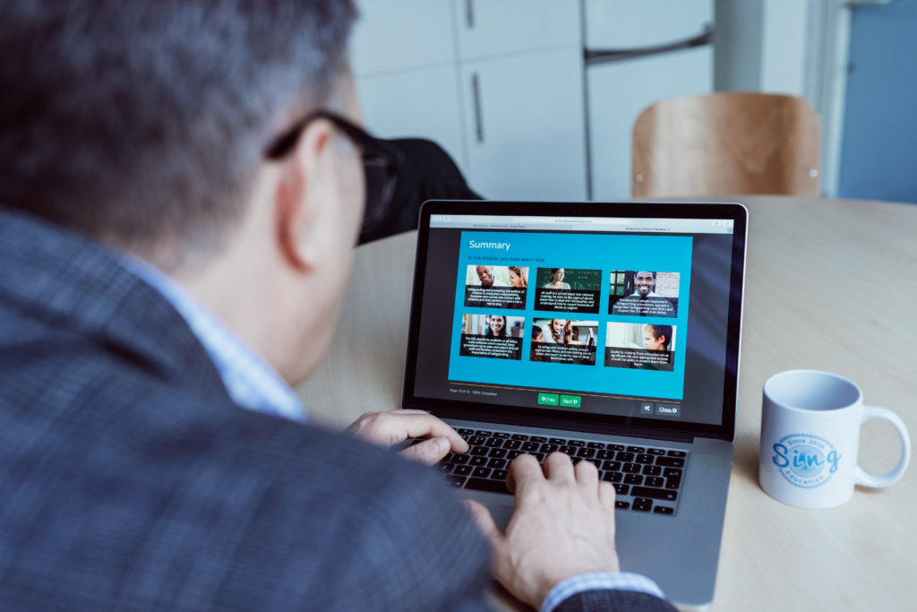 A professional is engaged with a laptop displaying a summary page with various text and image blocks, suggesting a learning or content management system. The person's focus on the screen, along with a coffee mug branded with "Sing Education" on the desk, conveys a setting of dedicated work or study, possibly related to educational planning or professional development.