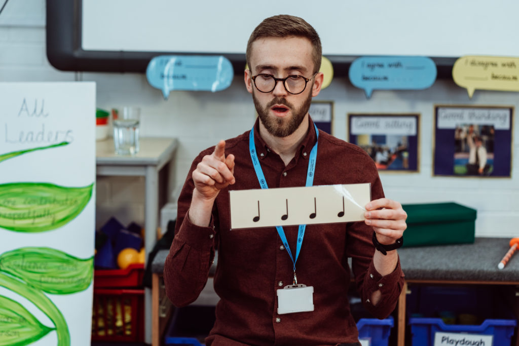 A focused educator with glasses and a beard, wearing a lanyard and ID badge, holds up a card with musical notes and gestures as if conducting or teaching rhythm. The classroom setting includes colorful educational posters and student work displayed on the walls, creating an environment of active learning.
