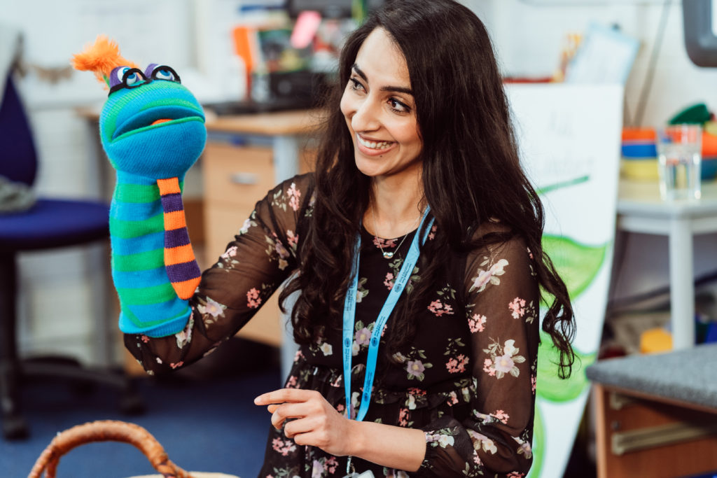 A smiling educator engages playfully with a colorful sock puppet, creating a lively and interactive learning environment. Her floral dress and the puppet's bright stripes and tuft of hair add a cheerful vibe to the classroom setting, filled with educational materials in the background, suggesting a focus on creativity and student involvement.