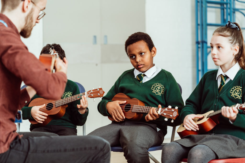 A music instructor is teaching ukulele to a small group of attentive students wearing school uniforms. The focus is on a young boy as he carefully watches the teacher's hand positioning, while a girl to his side also practices, illustrating a moment of musical learning and concentration.
