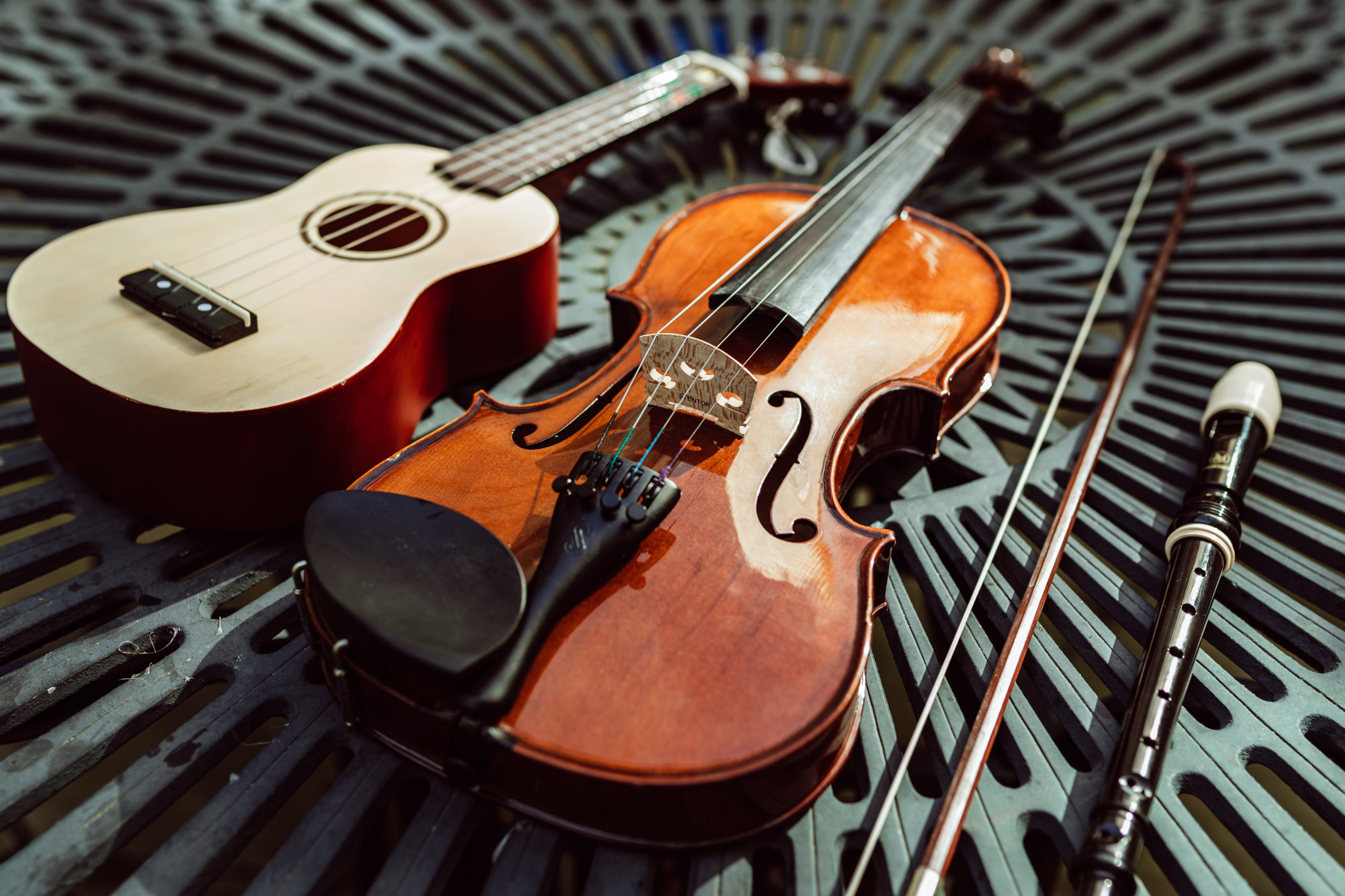 A violin with its bow and a ukulele are placed on a metal grate surface, bathed in warm sunlight, suggesting a setting for music practice or an outdoor performance.