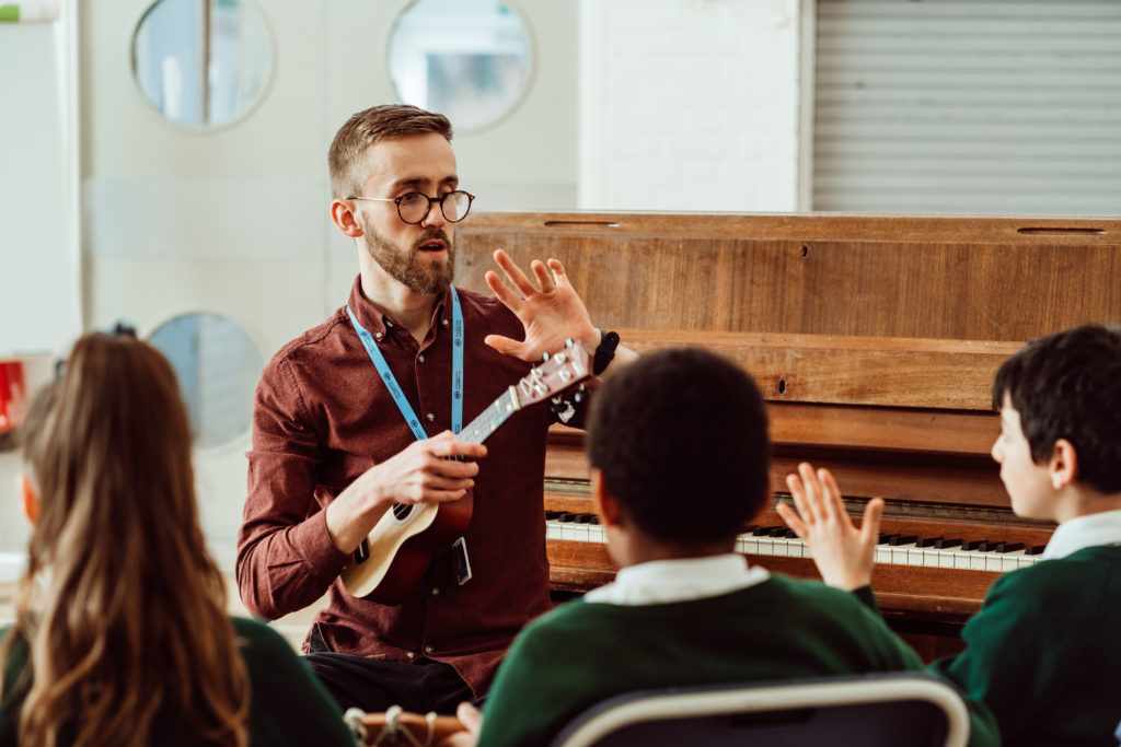 A music teacher, holding a ukulele, is engaging with young students in a classroom setting. The teacher is gesturing with his hand, possibly explaining a concept or giving instructions, while the students, wearing school uniforms, are attentively listening. Behind them, an upright piano suggests a hands-on music lesson is underway, fostering a creative and educational atmosphere.