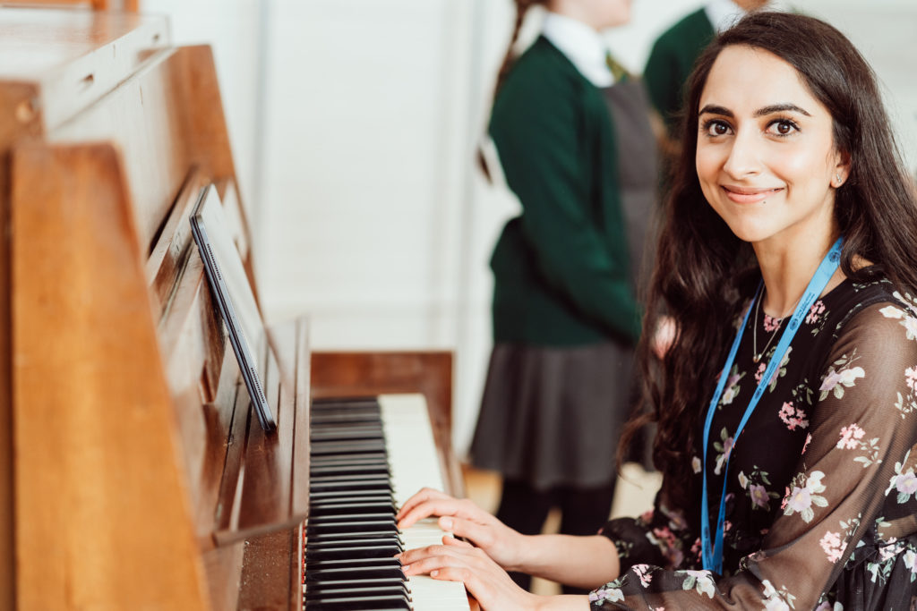 A smiling woman with a lanyard sits at a piano, her fingers poised on the keys, while a student in a green school uniform stands in the background, suggesting a music lesson in progress. The focus on the teacher's engaging expression conveys a sense of encouragement and the joy of sharing music.