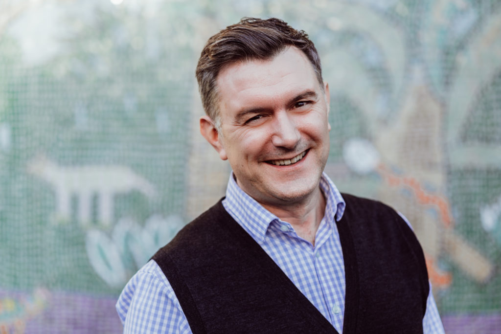A smiling man with a confident, friendly demeanor stands in front of a blurred mosaic background. He's dressed smartly in a blue checkered shirt with a dark vest, exuding a professional yet approachable vibe.
