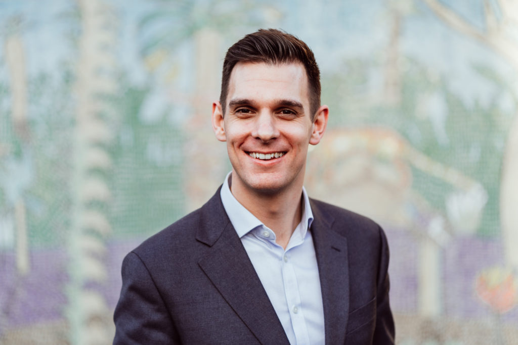A smiling man dressed in a smart grey suit jacket and a light blue shirt stands in front of a colorful mosaic background. His confident, friendly expression suggests a professional and approachable demeanor.