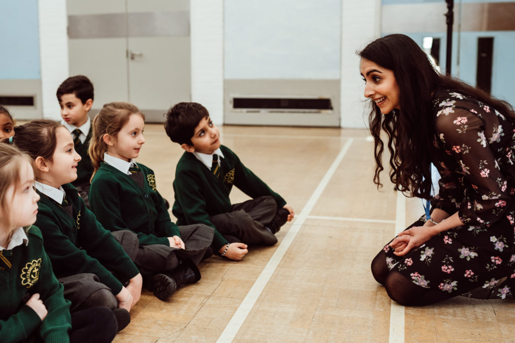 A group of young students in school uniforms sits attentively on the floor of a spacious room, facing a smiling teacher who is crouched down to their level. The teacher's engaging demeanor and the children's focused expressions suggest a dynamic and interactive learning environment. The atmosphere is one of warmth and attention, indicative of a positive educational experience.