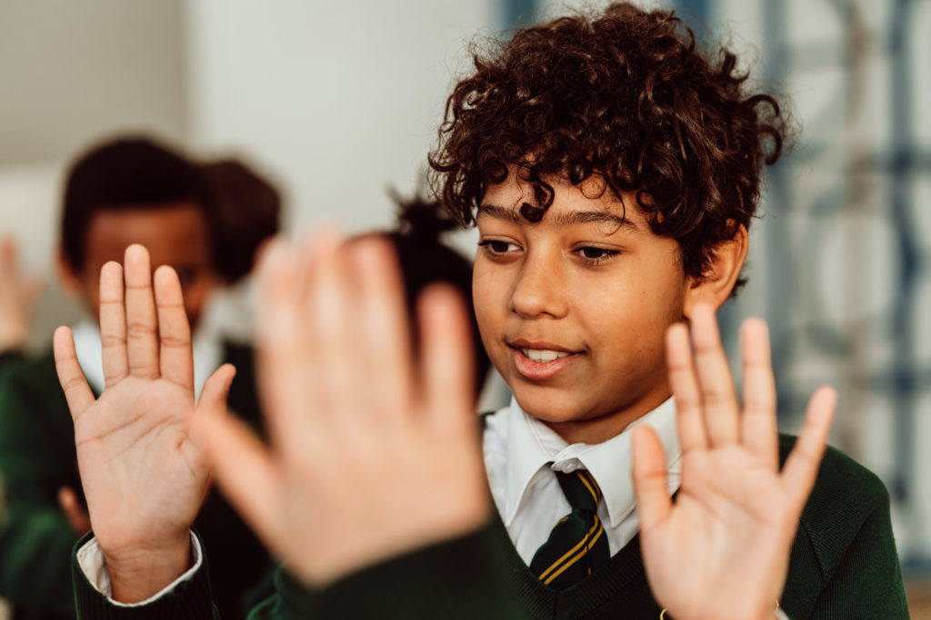 A young student with curly hair is actively participating in a classroom activity, raising their hands with a focused and joyful expression, while another student's hands are also visible in the foreground, suggesting a collaborative or interactive learning environment. The student is wearing a school uniform with a tie, indicating a formal educational setting.