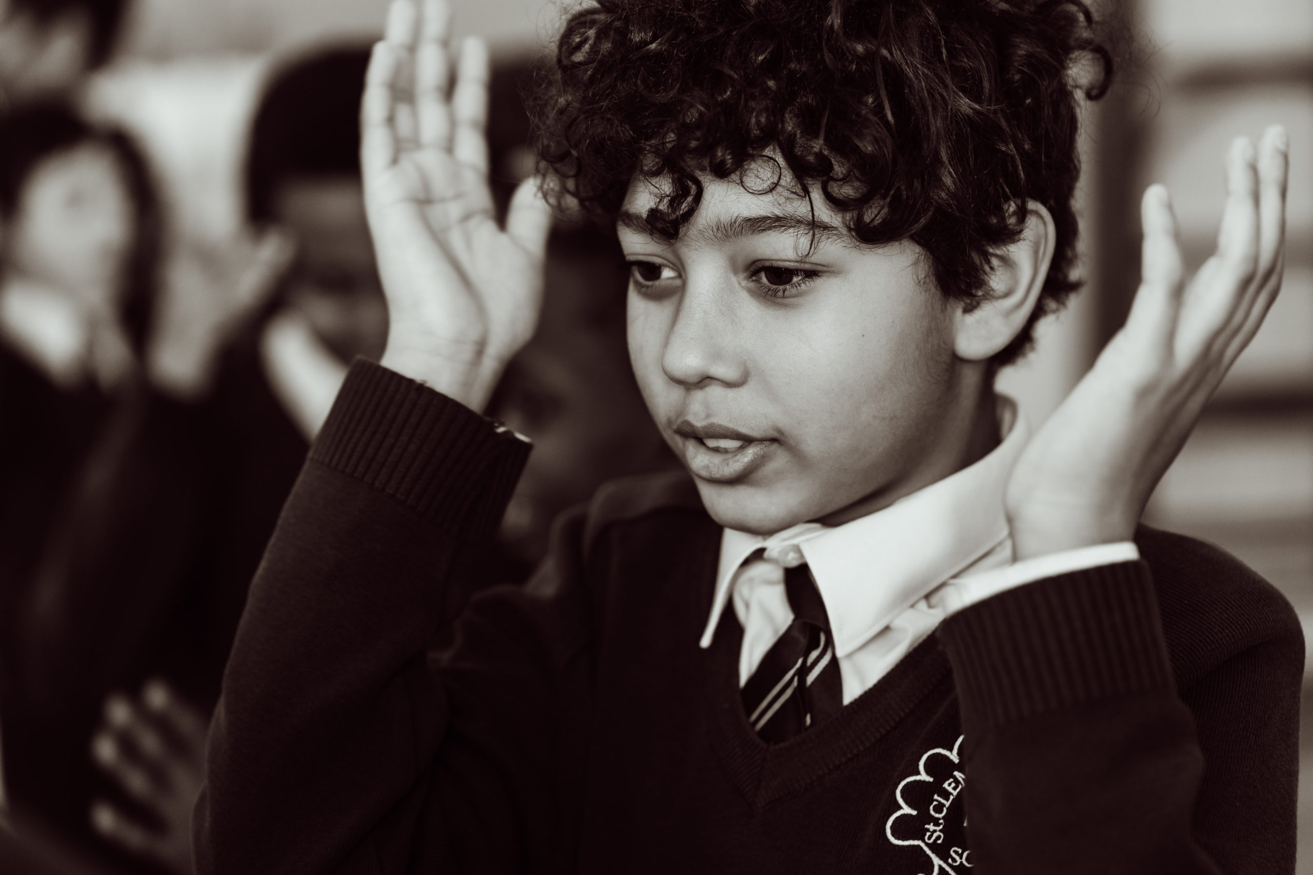 A young student with curly hair is captured in a moment of focus, hands raised near the sides of their face, possibly engaged in a classroom activity or responding to a teacher's instruction. The black and white photograph conveys a sense of concentration and participation, with other students slightly out of focus in the background, suggesting a group learning environment. The child is wearing a school uniform with a sweater that has the Sing Education logo, indicating an educational setting that fosters active involvement.