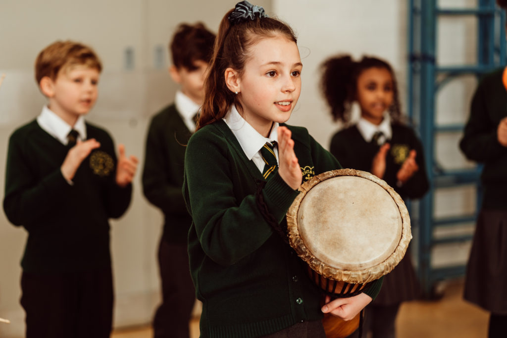 A group of schoolchildren in uniform are engaged in a music class, with one girl in the foreground confidently holding a drum, ready to play. Her classmates are clapping along, creating an atmosphere of collaborative learning and enjoyment in music education.