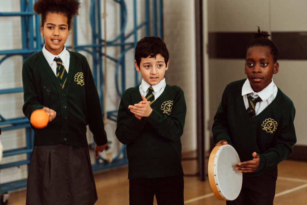 Three schoolchildren in green sweaters and striped ties participate in a music activity in a gymnasium, with one playing a tambourine, another holding a shaker, and the third clapping hands, all displaying focused expressions indicative of their engagement in the rhythmic exercise.