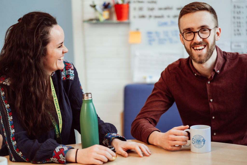 A man and a woman sit across from each other at a table, sharing a moment of laughter and connection. The woman, wearing a lanyard, is turned towards the man, her smile reflecting genuine engagement. The man, holding a mug with the Sing Education logo, reciprocates with a warm, friendly grin. The background hints at a casual, creative workspace with a whiteboard and colorful containers, suggesting a collaborative and relaxed environment.