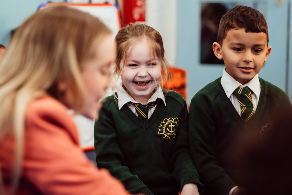 A group of young students, wearing green school uniforms with embroidered crests, share a joyful moment together. A girl in the center laughs heartily, her happiness infectious, while a boy to her right looks on with a gentle smile, and another student, partially out of focus, engages in the interaction. The warmth of the classroom setting suggests a lively and positive learning environment.