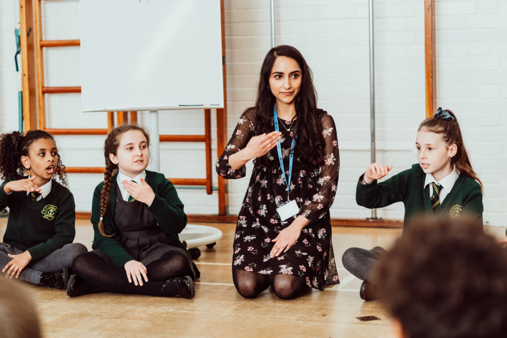 A music educator kneels among a group of attentive students seated on the floor, engaging them in a hand-signing activity. The children, wearing school uniforms, appear focused and animated, participating eagerly in the interactive learning experience. The atmosphere is one of active learning, with the whiteboard in the background suggesting a structured lesson in a bright classroom setting.