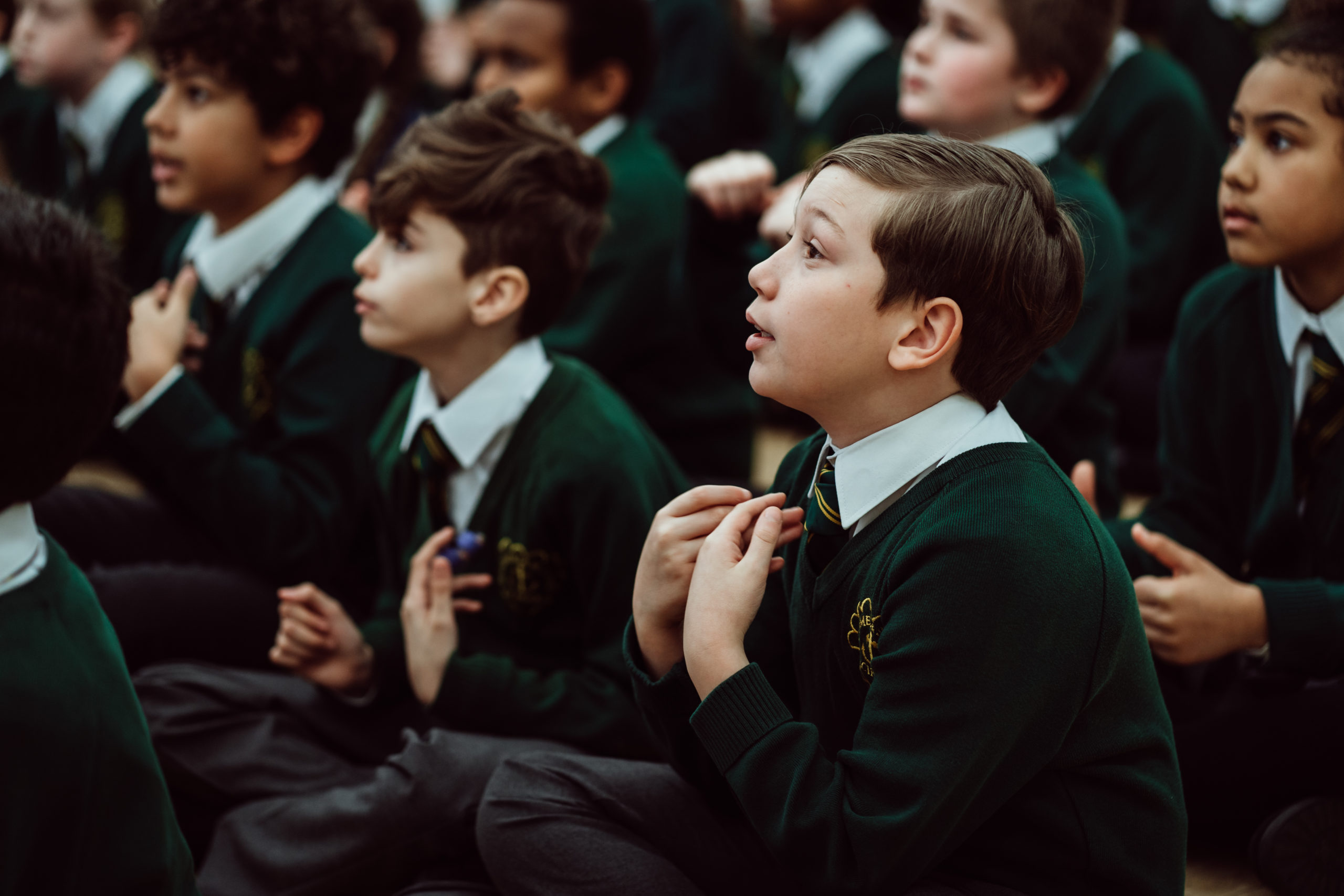 A group of schoolchildren, dressed in green uniforms with embroidered crests, sit attentively on the floor. Their expressions range from curiosity to engagement, with some children looking upwards, possibly at a teacher or during an assembly. The atmosphere suggests a moment of learning or listening, capturing the children's focus and interest in the activity at hand.