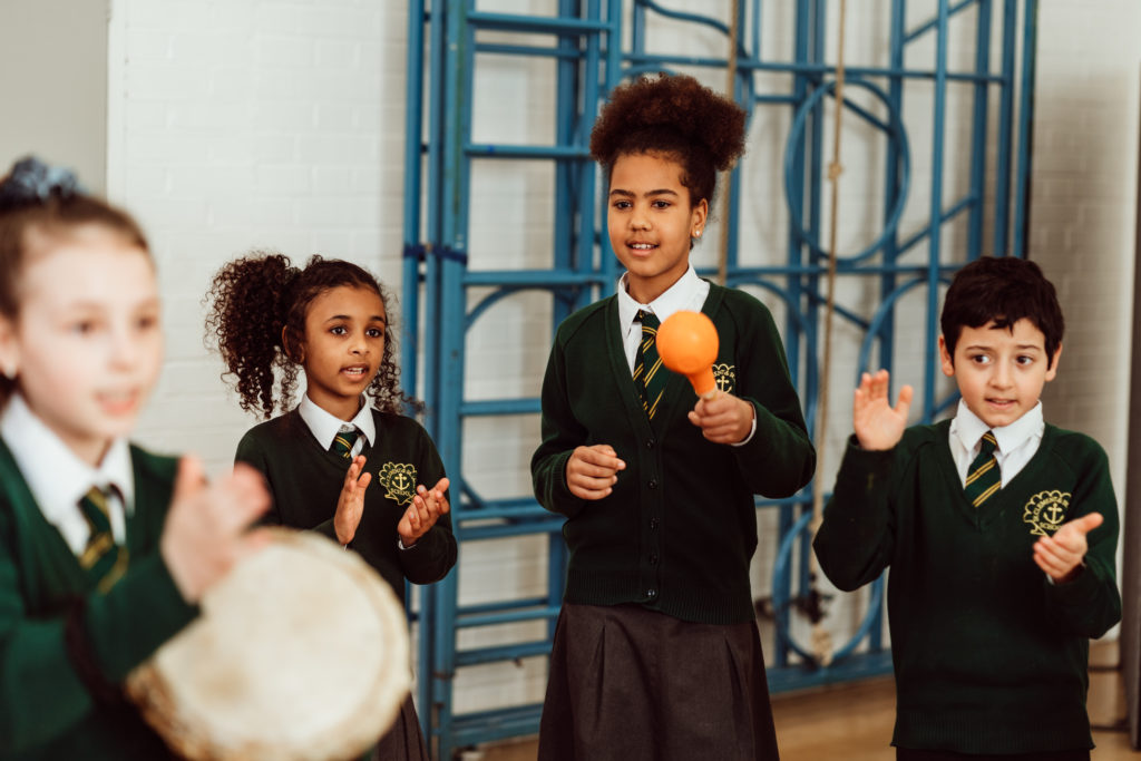 A group of schoolchildren in uniform are actively engaged in a music class, with one child in the foreground playing a tambourine and another confidently shaking a maraca, while their classmates participate with clapping and attentive expressions, all contributing to a lively and collaborative learning environment.