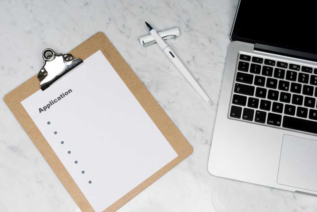 The image shows a modern workspace with a clipboard containing a blank application form, a white pen, and a laptop on a marble surface. The scene suggests a professional setting, possibly for job application or administrative work.