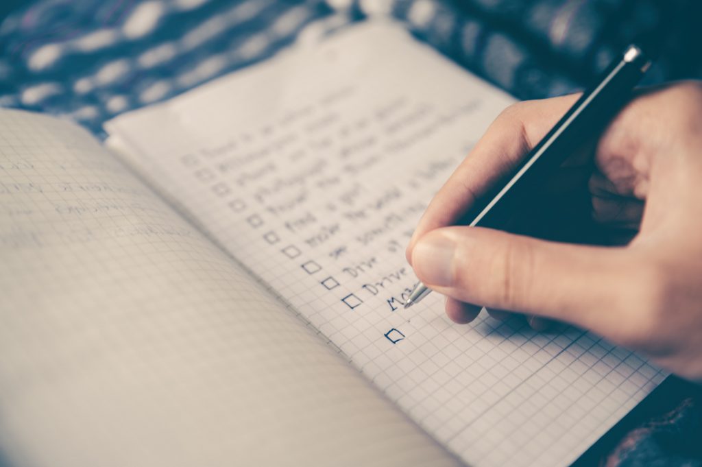 A person's hand is shown writing on a checklist with a pen, marking off an item on the list. The image captures the act of organizing or completing tasks, with a focus on the hand, pen, and paper, conveying a sense of productivity and task management.