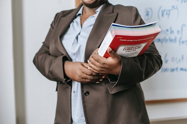 A professional individual stands in front of a whiteboard, holding a stack of textbooks and notebooks close to their chest. The person is wearing a brown blazer over a light blue shirt, and the whiteboard behind them features mathematical equations, suggesting an educational or academic setting. The focus on the books and the partial view of the individual implies a sense of readiness and commitment to learning or teaching.