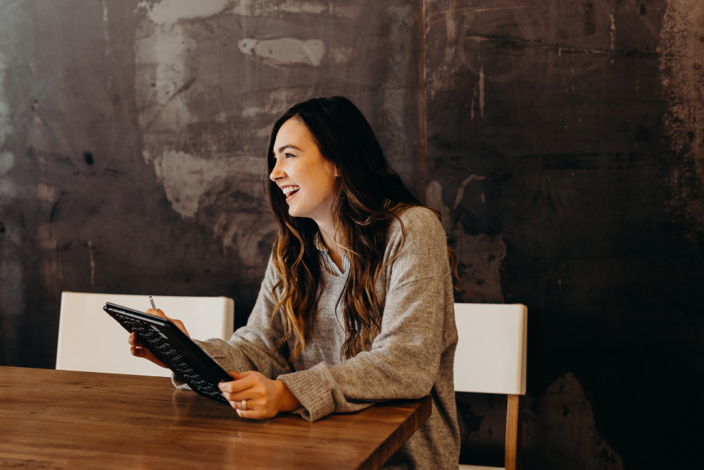 A smiling woman sits at a wooden table, holding a calculator and looking to the side with an expression of engagement and amusement. She's wearing a casual grey sweater, and the background features a textured dark wall, providing a contrast that highlights the warmth of the scene.