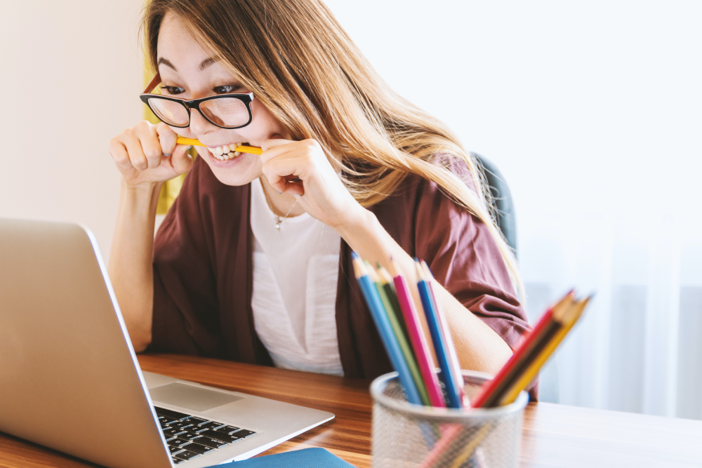 A young woman with glasses is biting a pencil while intensely focusing on the screen of her laptop. She's seated at a wooden desk with a cup filled with colorful pencils beside her, suggesting a setting of study or work. The atmosphere conveys concentration and perhaps the stress of meeting a deadline or solving a complex problem.