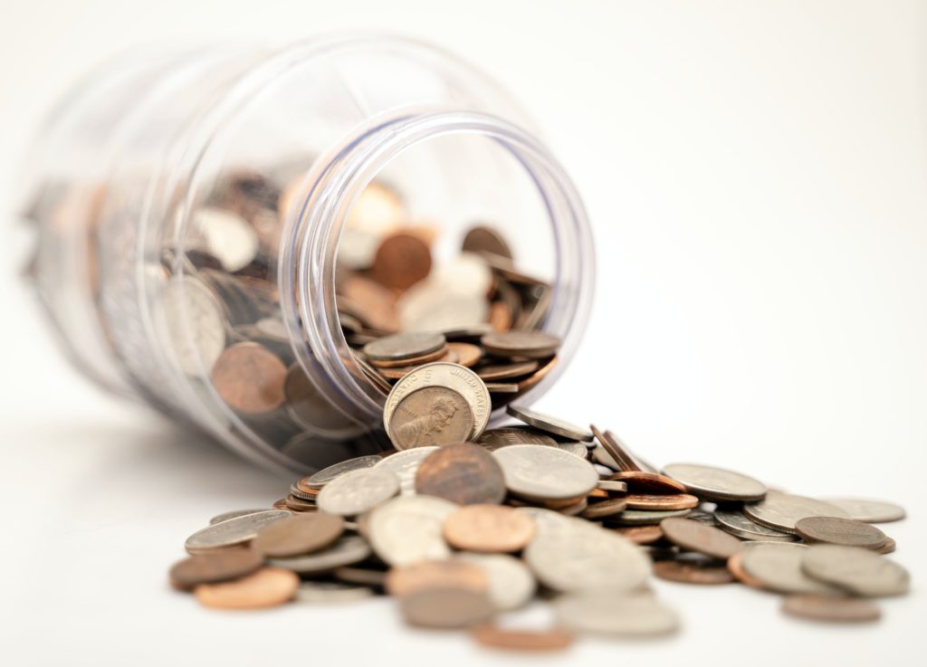 A clear jar tipped over with a multitude of coins spilling out onto a white surface, suggesting themes of savings, finance, or economy. The focus is on a coin in the foreground, with the rest of the image softly blurred.