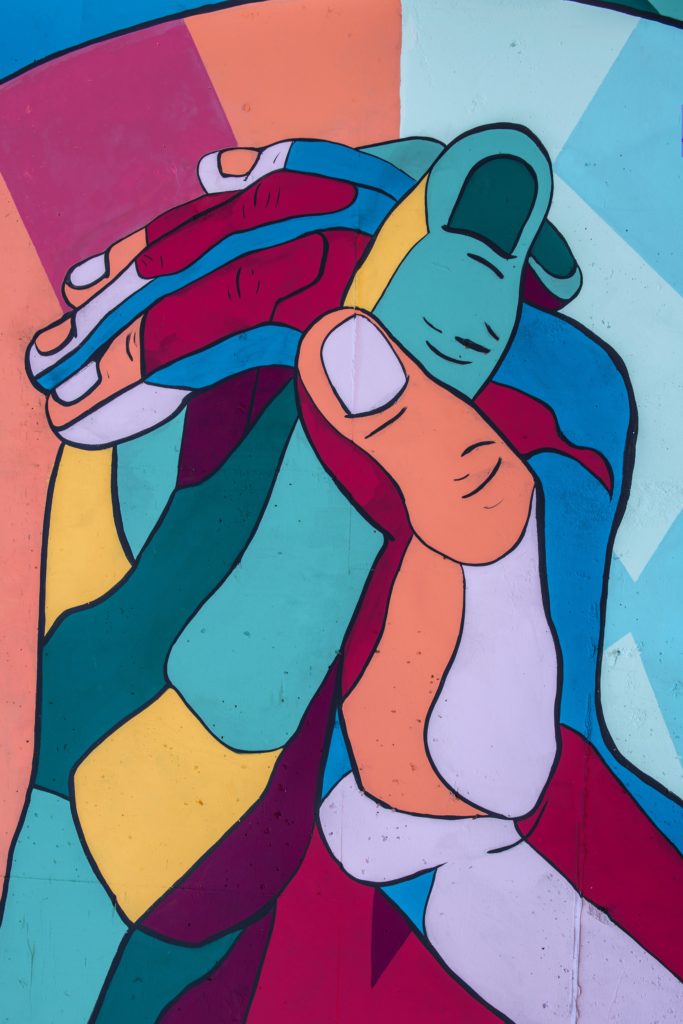 The image displays a vibrant and colorful mural of interlocking hands. The hands are depicted in a variety of bold colors, with outlines and shading that give the impression of movement and unity. The background consists of geometric shapes in contrasting colors, enhancing the visual impact of the artwork.