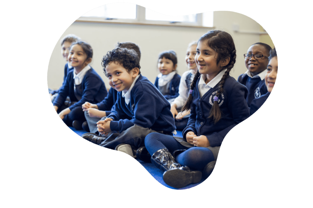 A group of joyful schoolchildren, dressed in blue uniforms, are seated on the floor, their attention seemingly captured by an engaging activity. Their bright smiles and animated expressions convey a sense of excitement and camaraderie, reflecting the lively and positive atmosphere of a classroom environment.