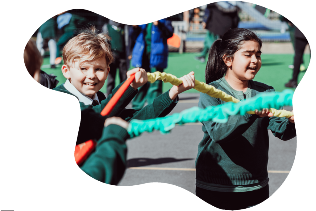 Two children are engaged in a playful tug-of-war game, gripping a colorful rope with determination and joy. The boy on the left smiles broadly, while the girl on the right appears focused and spirited, both wearing green school uniforms. The outdoor setting and their classmates in the background suggest a lively schoolyard atmosphere.