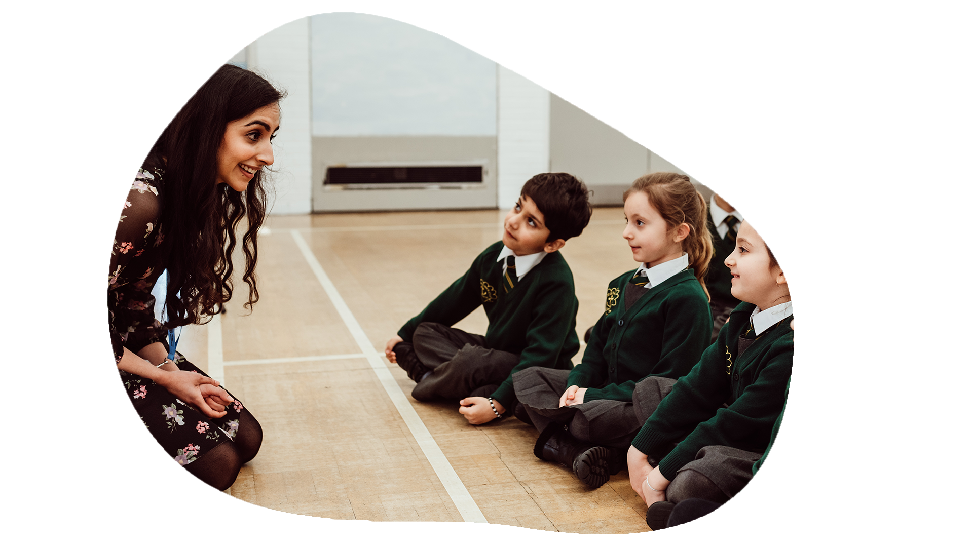 A smiling teacher in a floral dress kneels to engage with three attentive students sitting cross-legged on the floor, all wearing school uniforms, in a bright classroom setting, fostering a warm and interactive learning environment.
