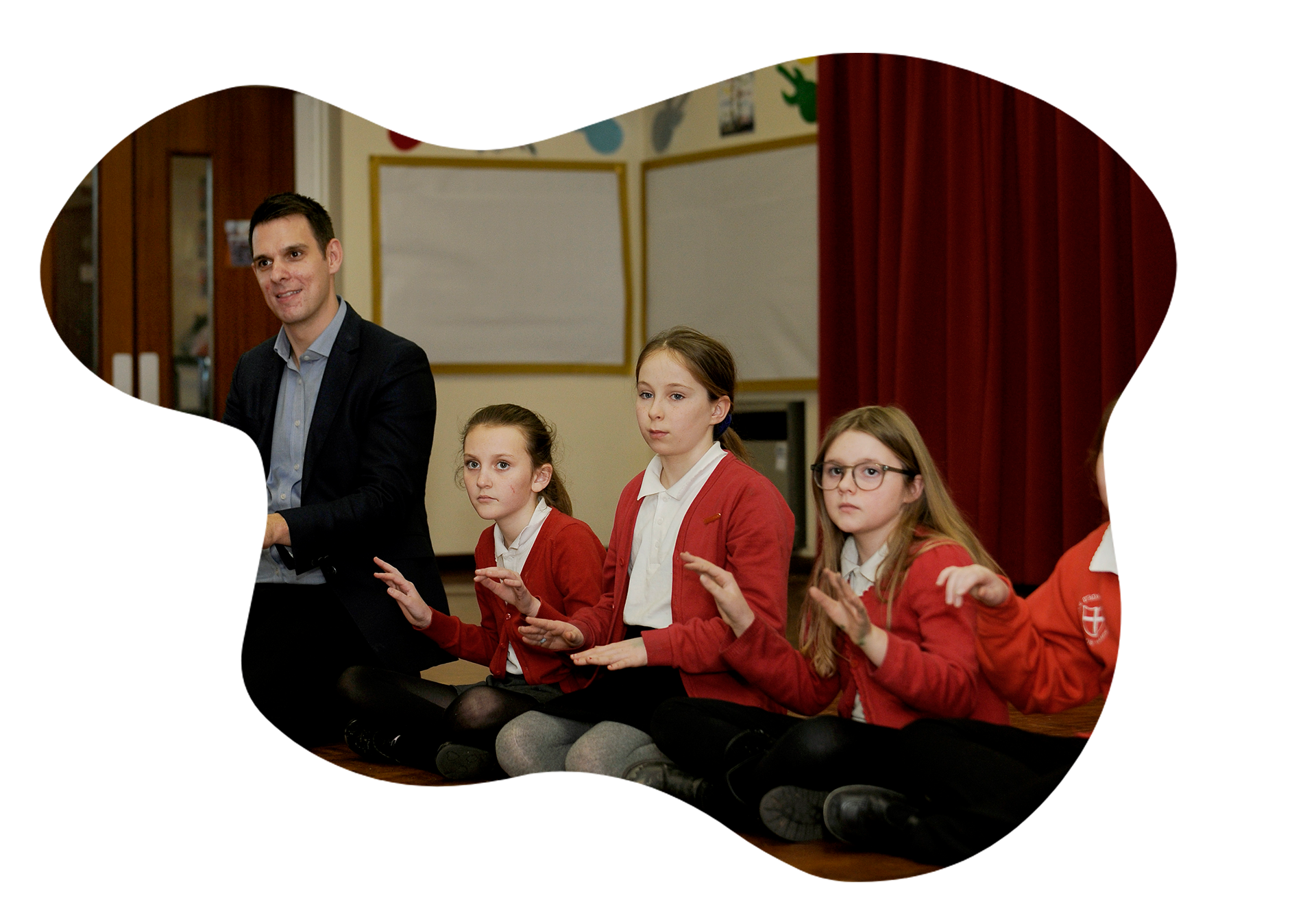 A group of focused students in red uniforms and a teacher are seated on the floor, practicing hand percussion with visible engagement and concentration, in a classroom adorned with colorful handprints on the wall and a red curtain backdrop, creating an atmosphere of interactive learning.