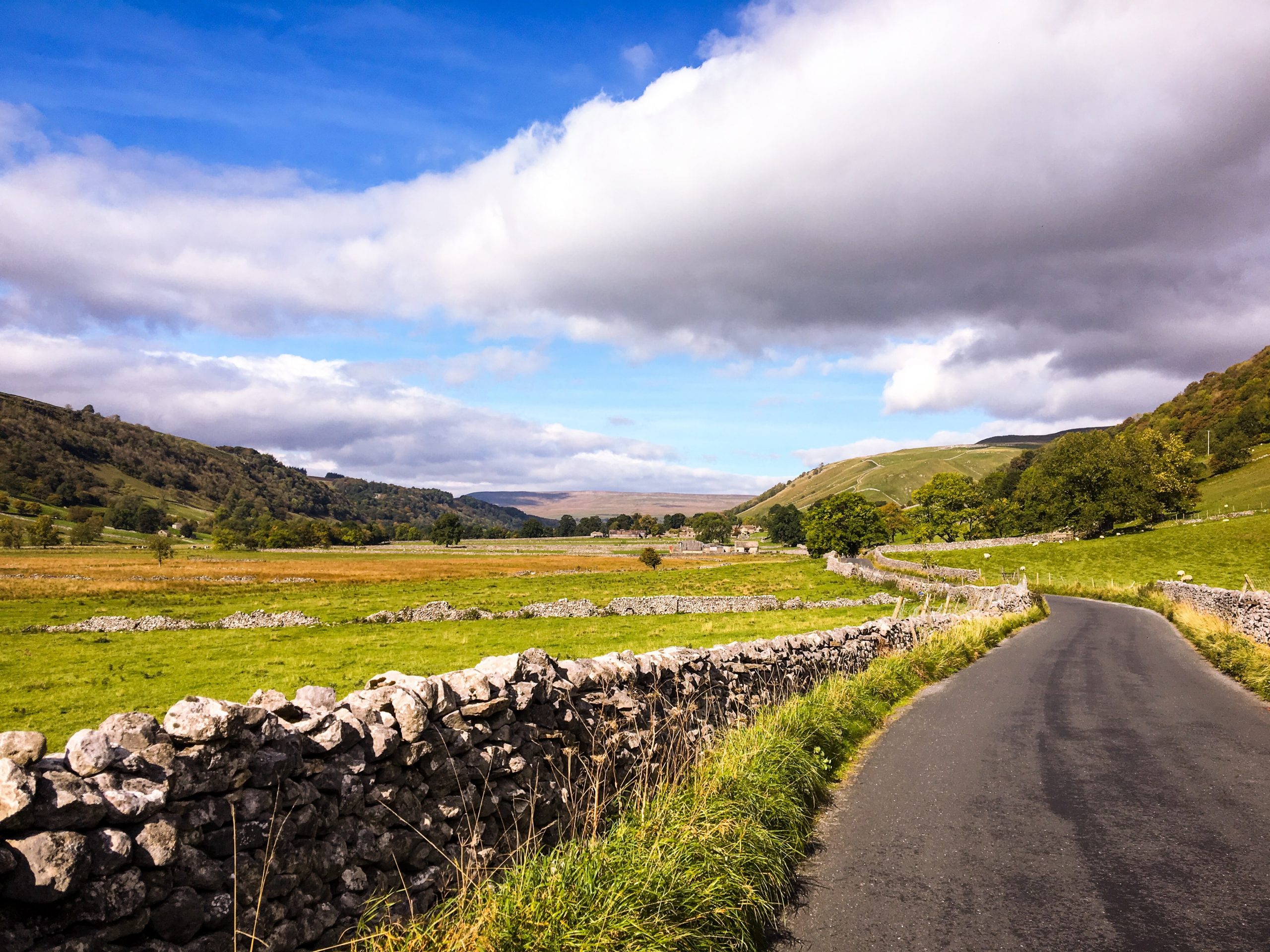 A winding country road leads through a picturesque rural landscape, flanked by traditional stone walls and expansive fields. Rolling hills rise in the distance under a dynamic sky, half-filled with fluffy white clouds and the other half with darker, more ominous ones, suggesting a changeable weather typical of a lush, verdant countryside.