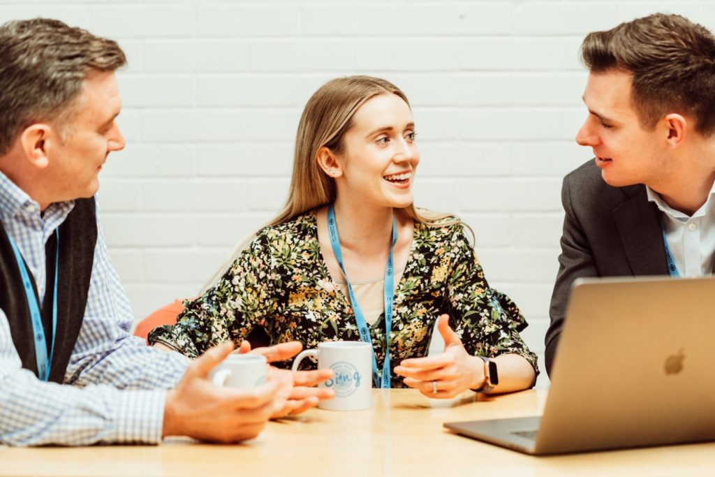 Three professionals are engaged in a lively discussion around a table, with a laptop open in front of them. The woman in the center, wearing a floral top and a lanyard, is animatedly talking to the man on the right, who is attentively listening and wearing a suit. The man on the left, also with a lanyard, is holding a coffee mug and appears to be part of the conversation, contributing to a collaborative and congenial atmosphere.