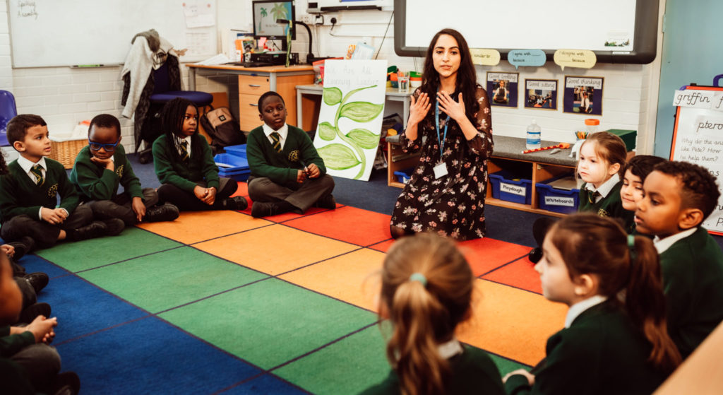 A teacher is engaging with young students seated on a colorful classroom rug. The attentive children, dressed in green uniforms, watch as the teacher gestures expressively, fostering a lively and interactive learning environment. The backdrop of the classroom is filled with educational materials and student work, creating a vibrant and nurturing educational setting.