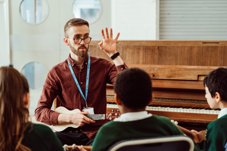 A music teacher with a beard and glasses is engaging with young students while holding a ukulele, raising his hand as if to illustrate a point or direct the class. The students, wearing green school uniforms, are attentively listening to the teacher, with the backdrop of a classroom featuring a piano, suggesting a hands-on music learning environment. The atmosphere conveys a sense of interactive learning and musical instruction.
