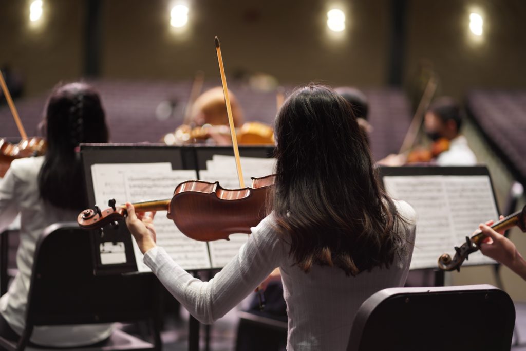 A group of violinists is captured from behind while performing, their bows raised in harmony. The focus is on a musician in the foreground, with sheet music on stands in front of them, and the blurred auditorium seats in the background suggest a rehearsal setting. The image conveys a sense of concentration and collaboration among the ensemble members.