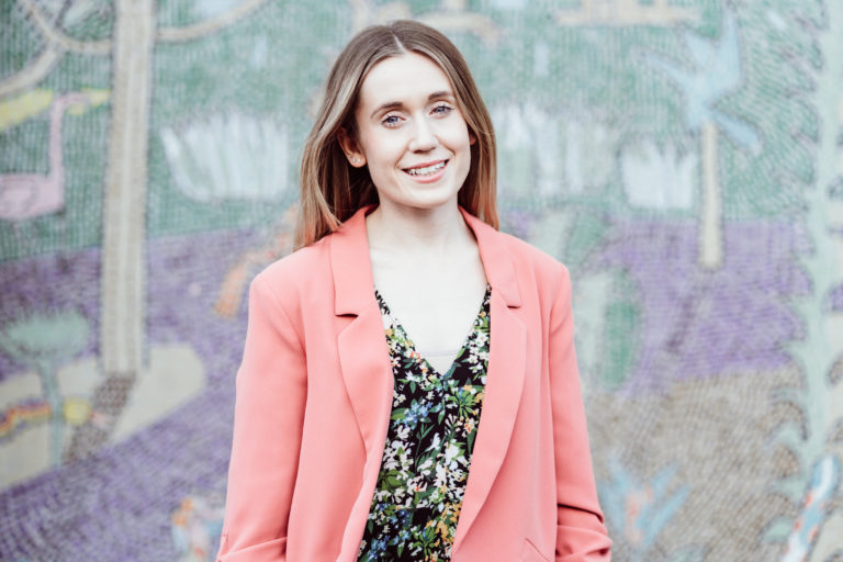A smiling woman with long hair wearing a coral blazer over a floral dress stands in front of a colorful mosaic wall. Her cheerful expression and casual pose convey a sense of approachability and confidence.