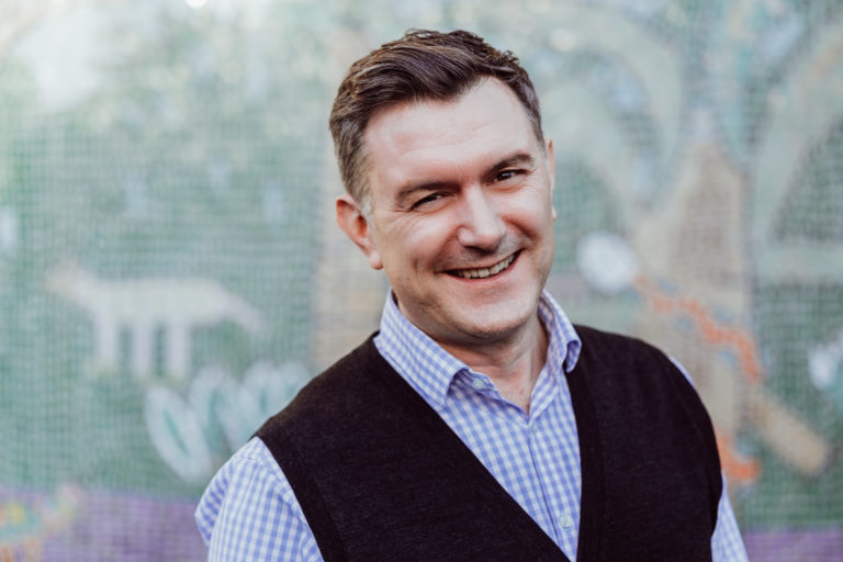 A smiling man wearing a checkered shirt and a dark vest stands in front of a colorful mosaic background. His confident, friendly expression suggests a welcoming and positive atmosphere.