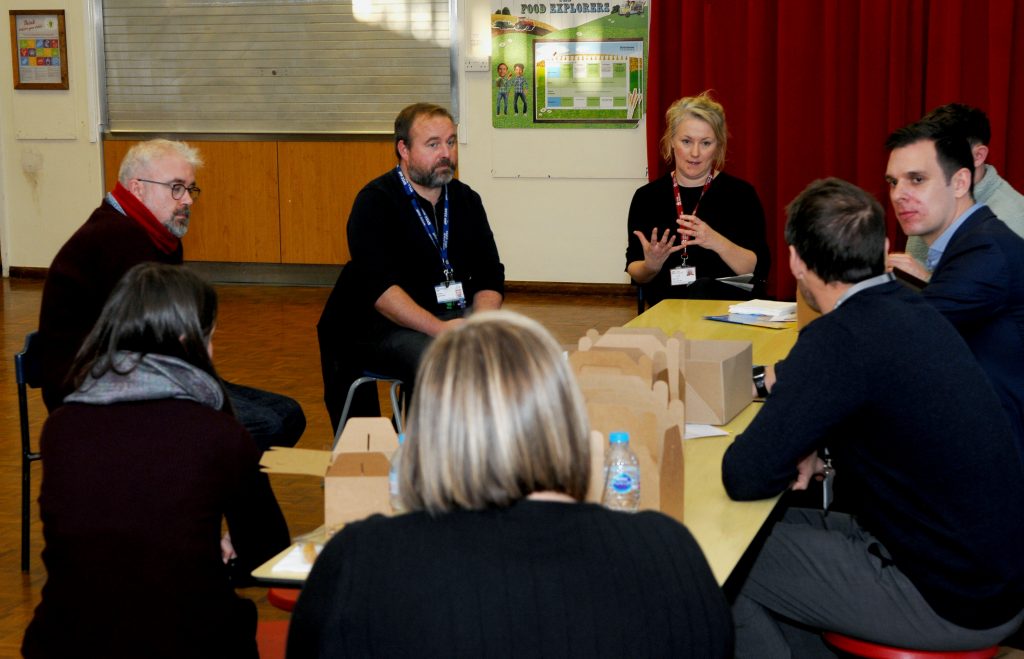 A group of professionals is engaged in a discussion around a table in a well-lit room. They appear focused and attentive, with some participants gesturing as they speak, indicating an active exchange of ideas. Open boxes of food suggest a working lunch environment, and the casual yet professional atmosphere hints at a collaborative meeting or training session.