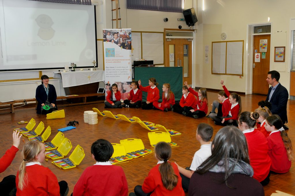 A lively music class unfolds in a spacious school hall where a group of students, clad in red uniforms, are seated on the floor, attentively participating in a lesson. Two adults, one playing a ukulele, guide the session. The room is filled with colorful boomwhackers and percussion instruments, suggesting an interactive and hands-on learning experience. A projector screen displays the title "Lemon Lime," hinting at a song or activity theme, while educational banners add context to the enriching environment.