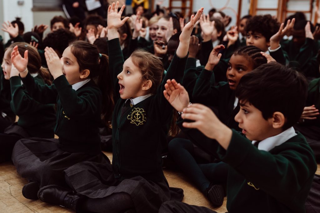A group of enthusiastic schoolchildren, clad in green uniforms with school crests, are seated on the floor, actively participating in a group activity. Their expressions are animated and joyful as they raise their hands and clap, fully engaged in the interactive learning experience. The atmosphere is lively and collaborative, highlighting the children's enjoyment and involvement in the educational moment.